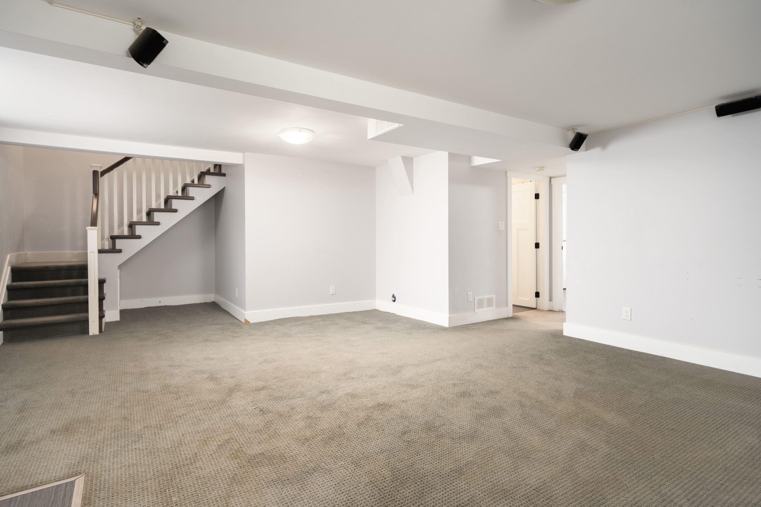 Empty basement room with carpet flooring, white walls, ceiling lights, and a staircase leading upstairs. A hallway and two doors are visible in the background.