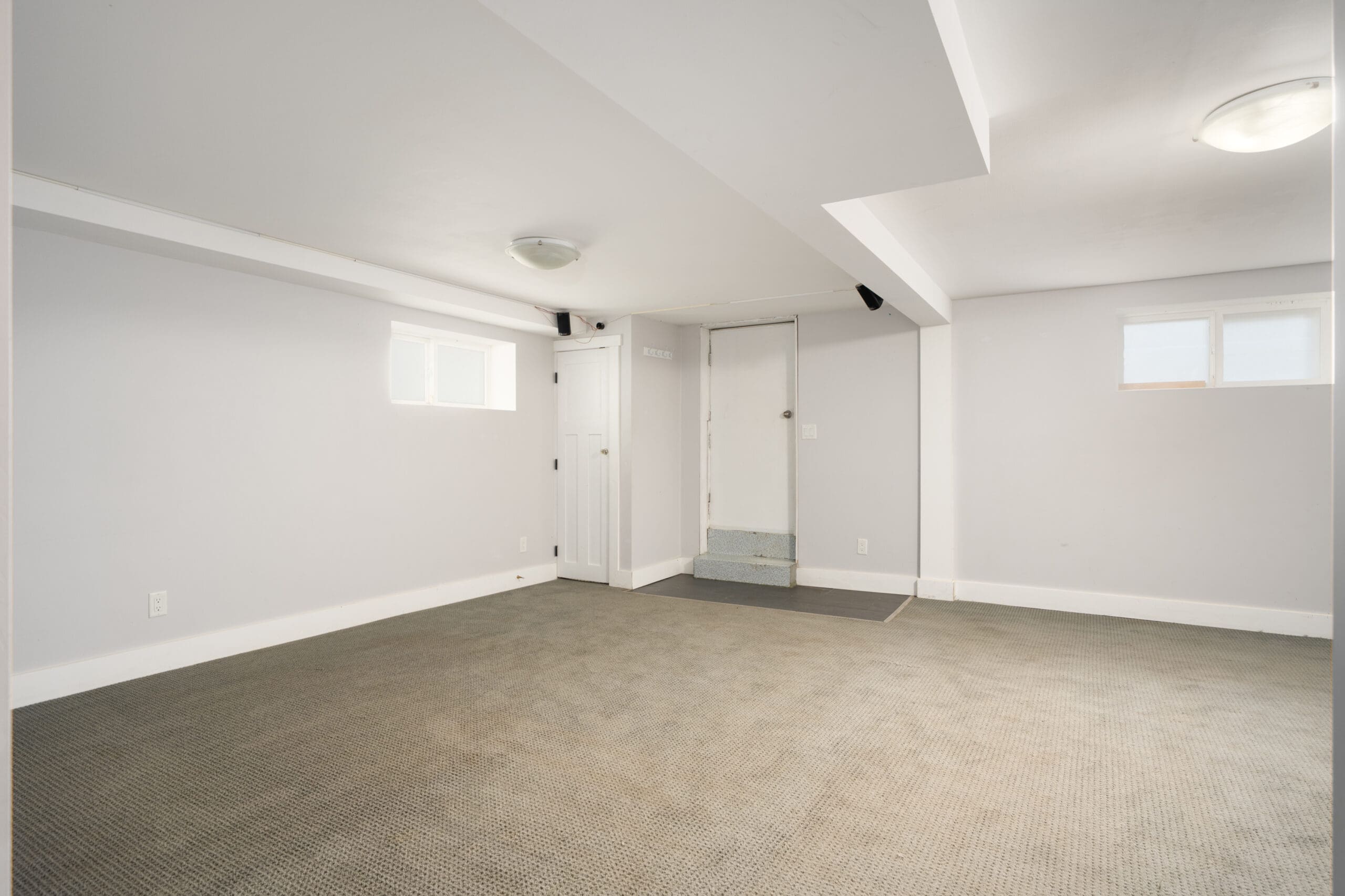 Unfurnished basement room with gray carpet, white walls, small windows, a door, and a few steps leading up to the entryway.