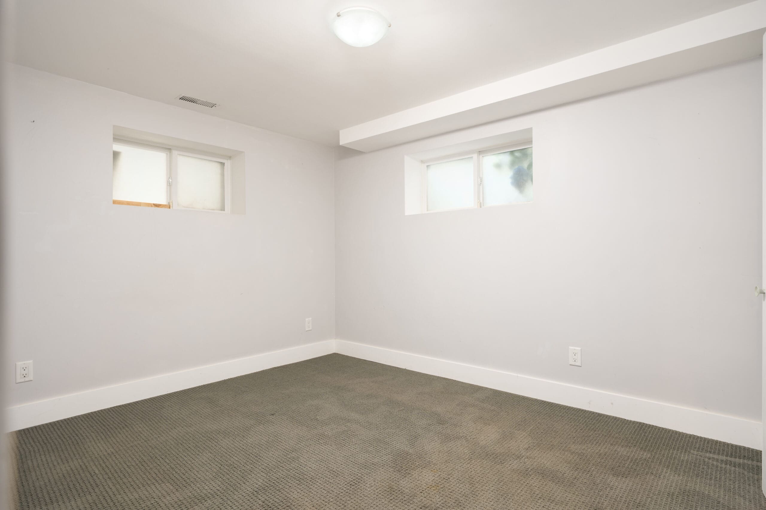 Empty room with light gray walls, two small rectangular windows, a ceiling light, and dark carpet flooring. No furniture or decor is present.