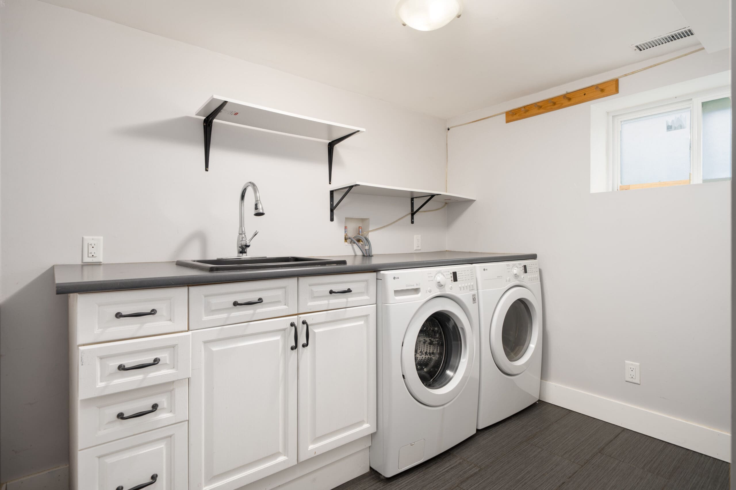 Laundry room with white cabinets, a countertop sink, shelves, a front-loading washing machine, and a dryer below a small window.