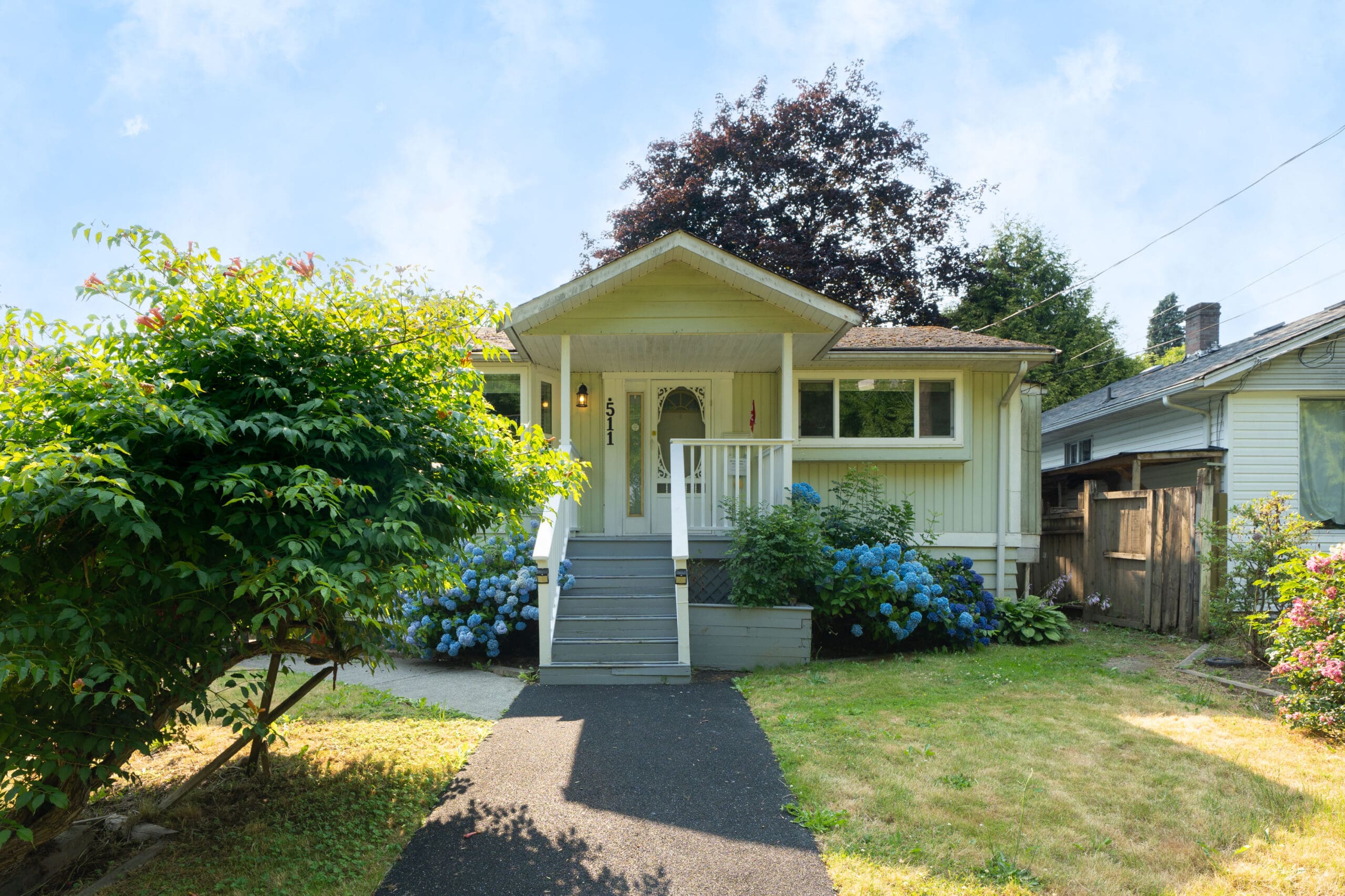 Single-story house with a covered porch, gray steps, and blue hydrangea bushes in front, surrounded by greenery under a clear sky.