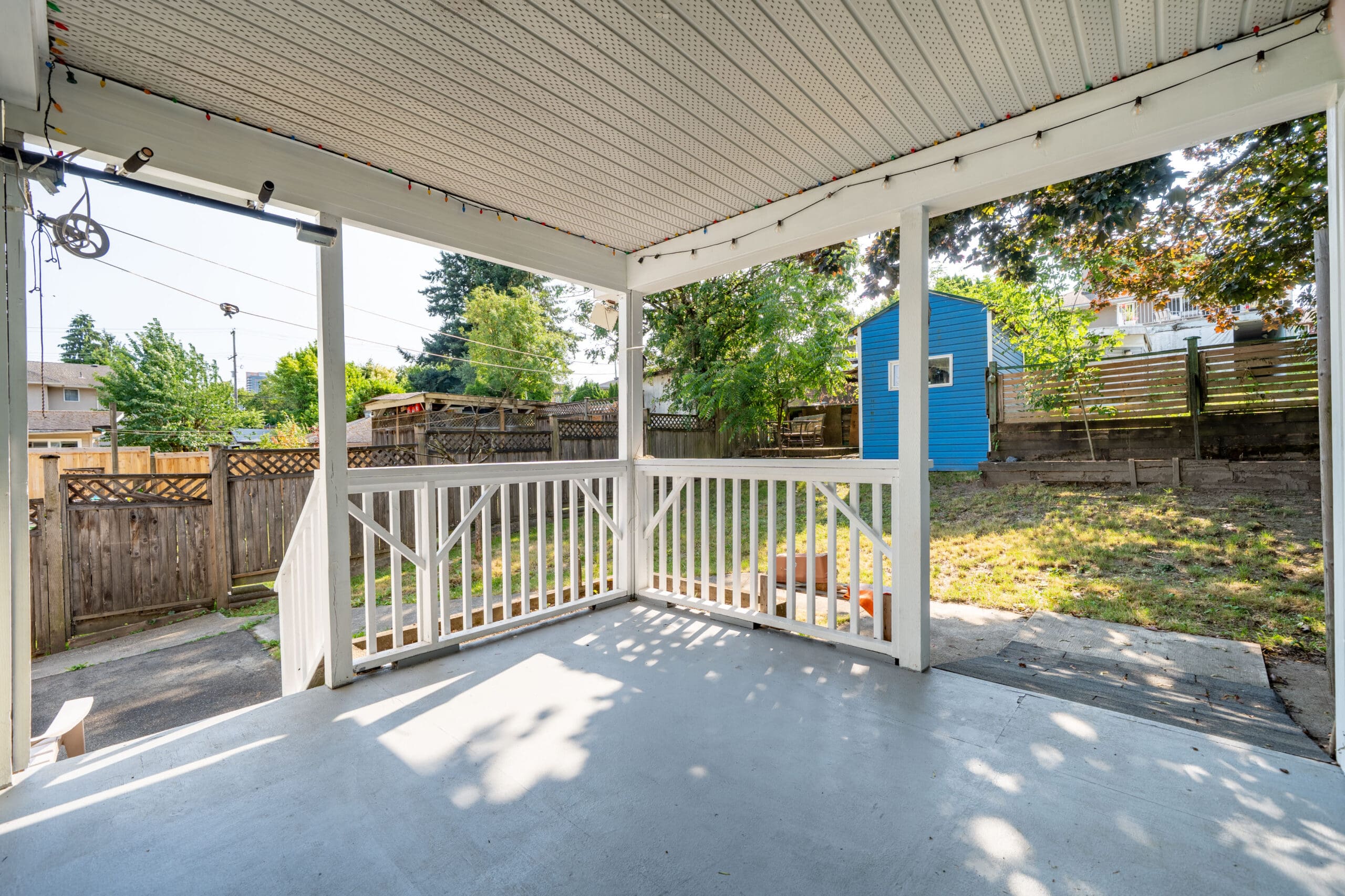 Covered outdoor patio with white railings overlooking a backyard with grass, trees, a blue shed, and a wooden fence on a sunny day.