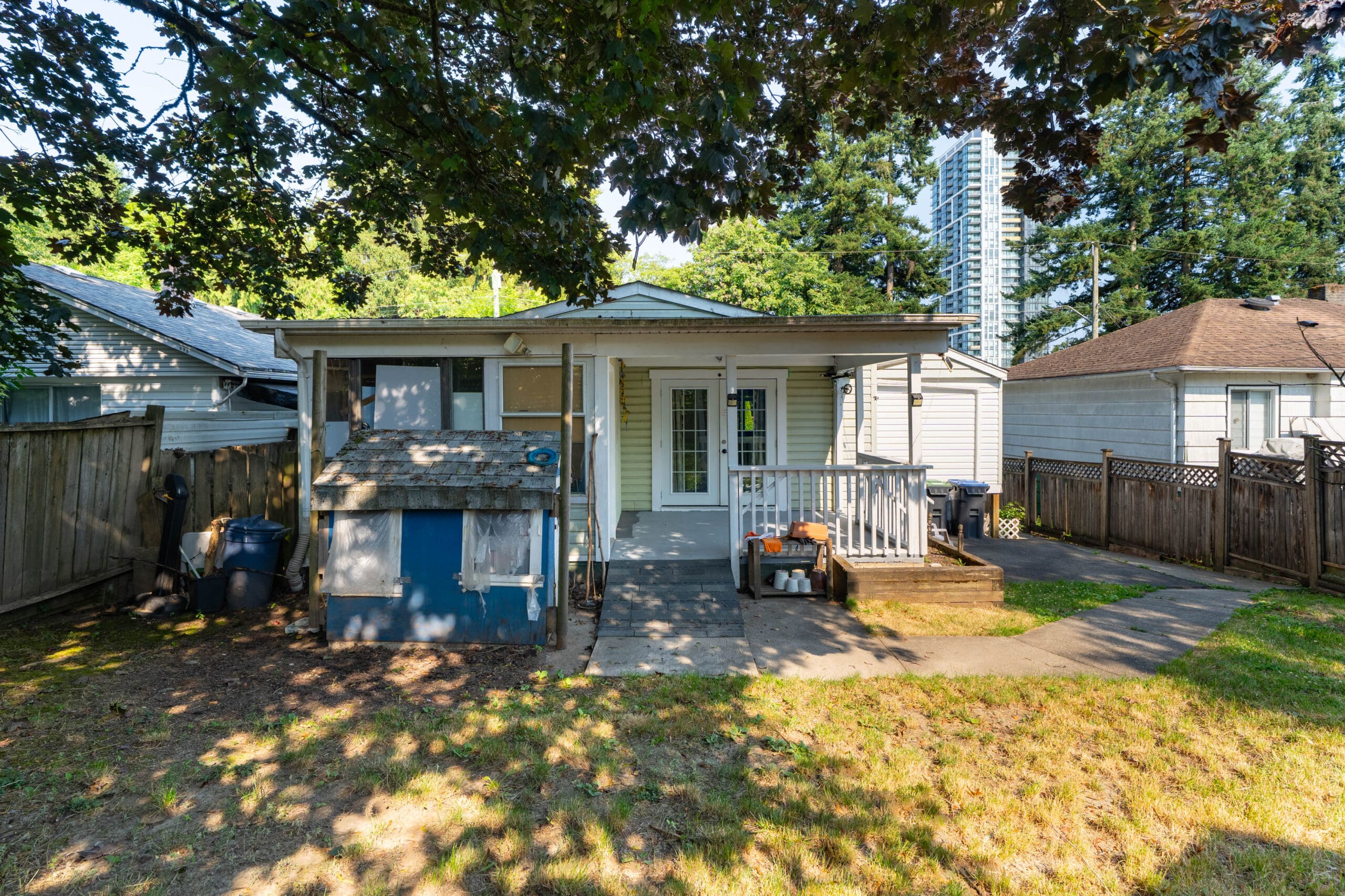 Small backyard with a covered patio, steps leading to a white back door, a blue shed, and fenced sides. Grass and trees are in the foreground, with neighboring houses visible.
