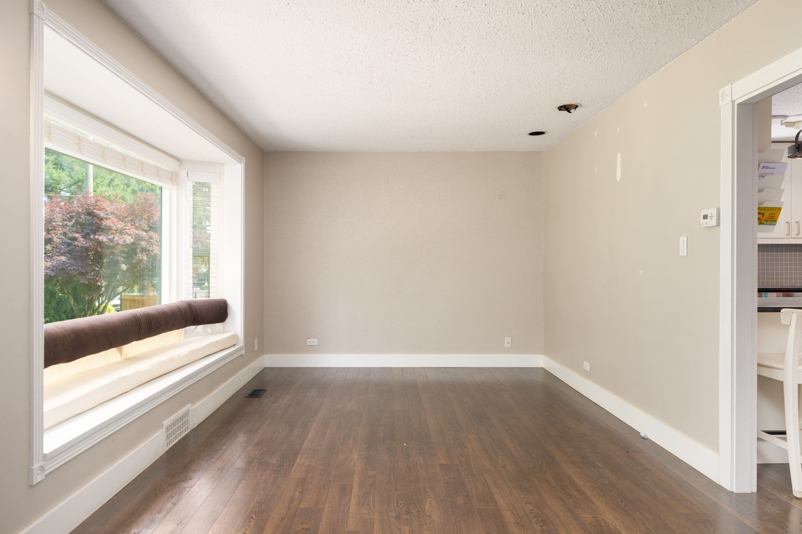 Empty room with light beige walls, dark wood flooring, a large bay window on the left, and an open doorway on the right leading to a white kitchen.