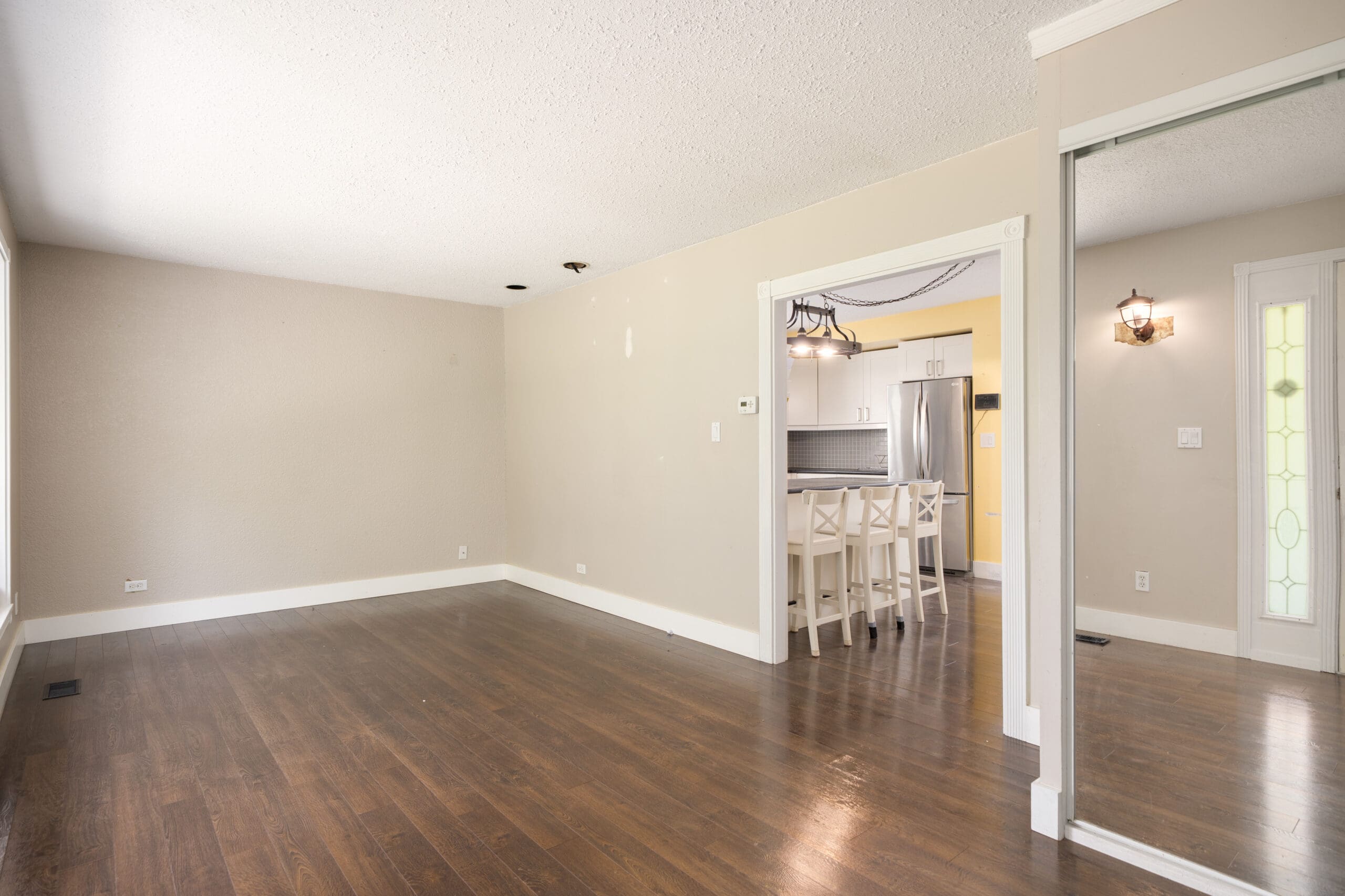 Empty living room with hardwood floor, beige walls, and large windows; open doorway leads to a kitchen with white cabinets and bar stools; mirror and front door visible on the right.