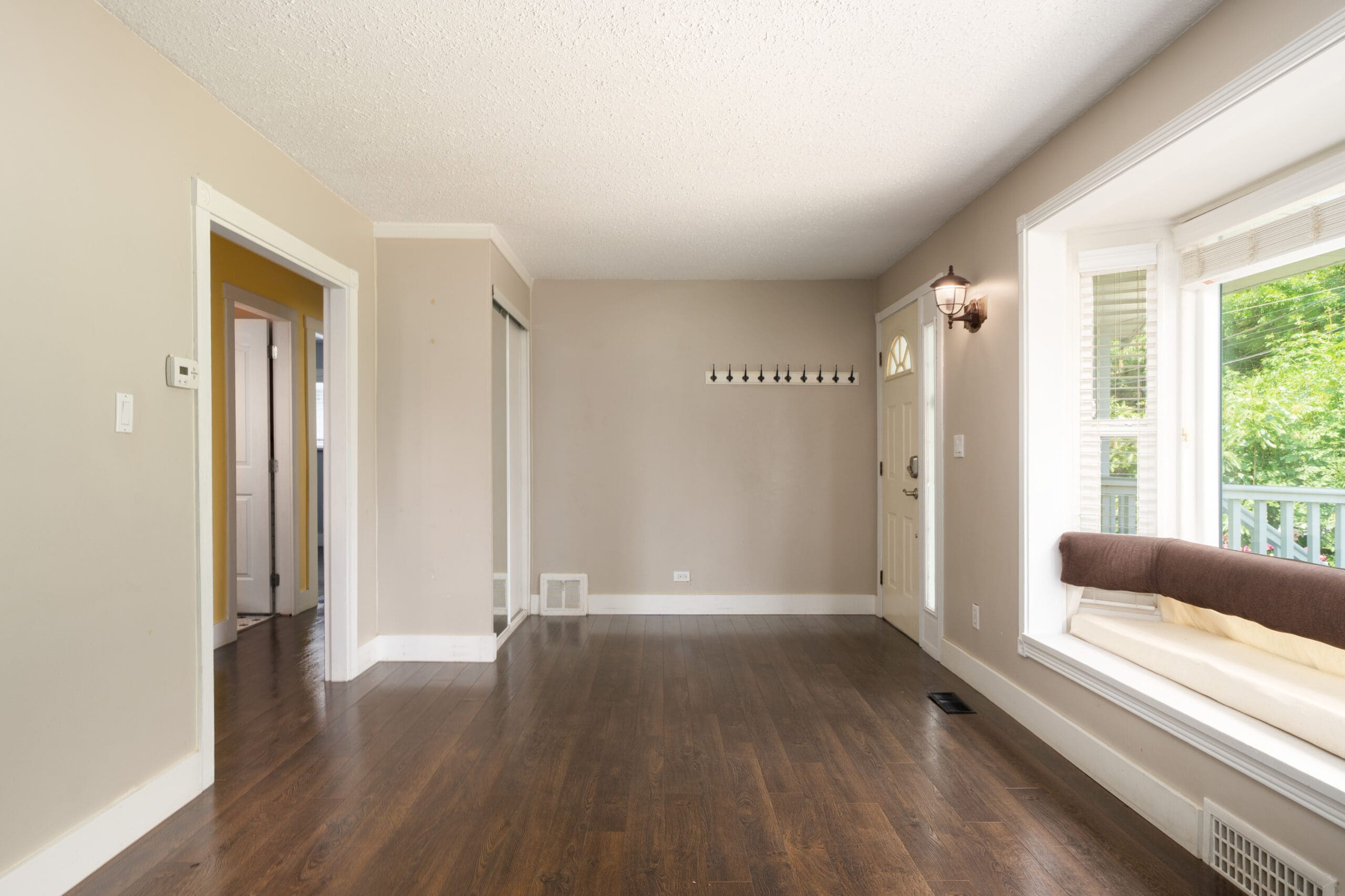 Empty living room with hardwood floors, beige walls, a front door, coat hooks, window seat, and large window letting in natural light.