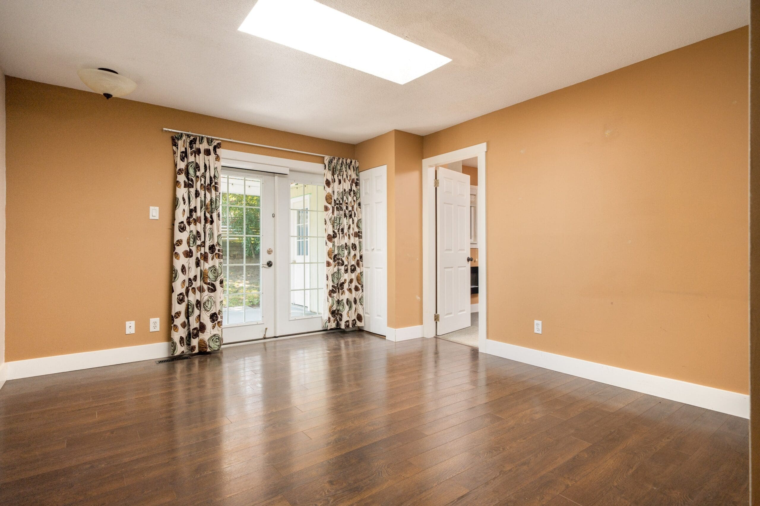 Empty room with tan walls, dark wood floor, a skylight, patterned curtains on glass doors, and an open white door.