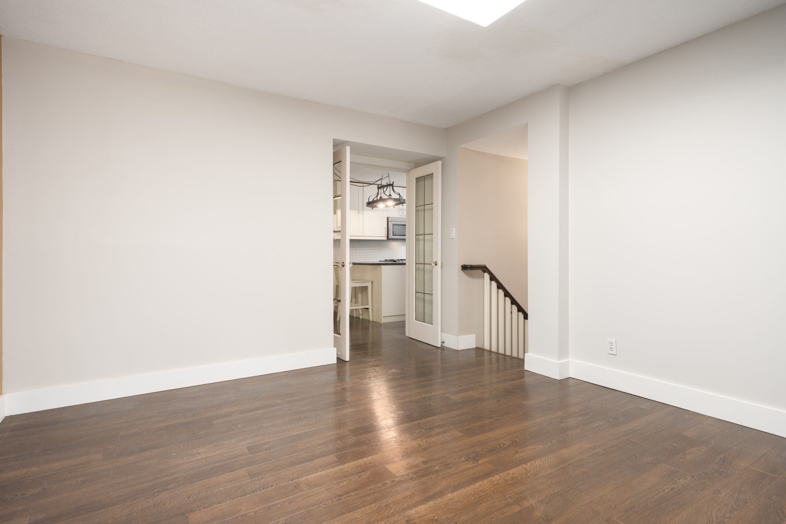 Empty room with hardwood floors, beige walls, and double glass doors leading to a kitchen area; staircase visible to the right.