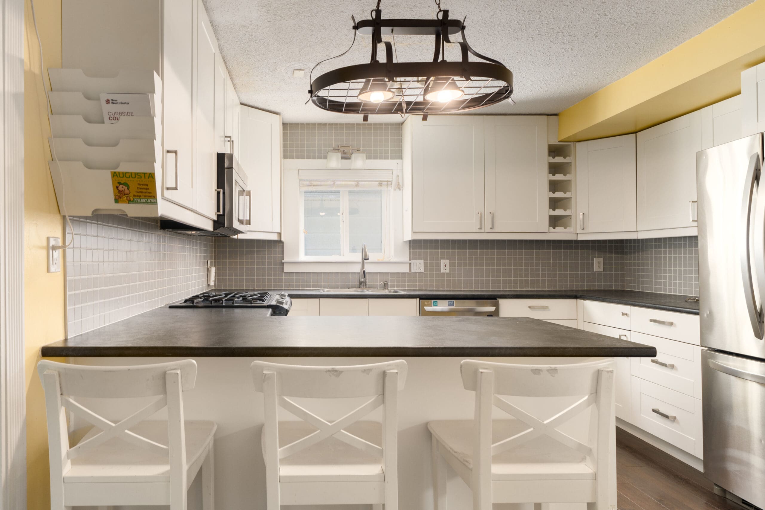 Modern kitchen with white cabinets, black countertop island, three white chairs, stainless steel appliances, and a large industrial-style pendant light overhead.