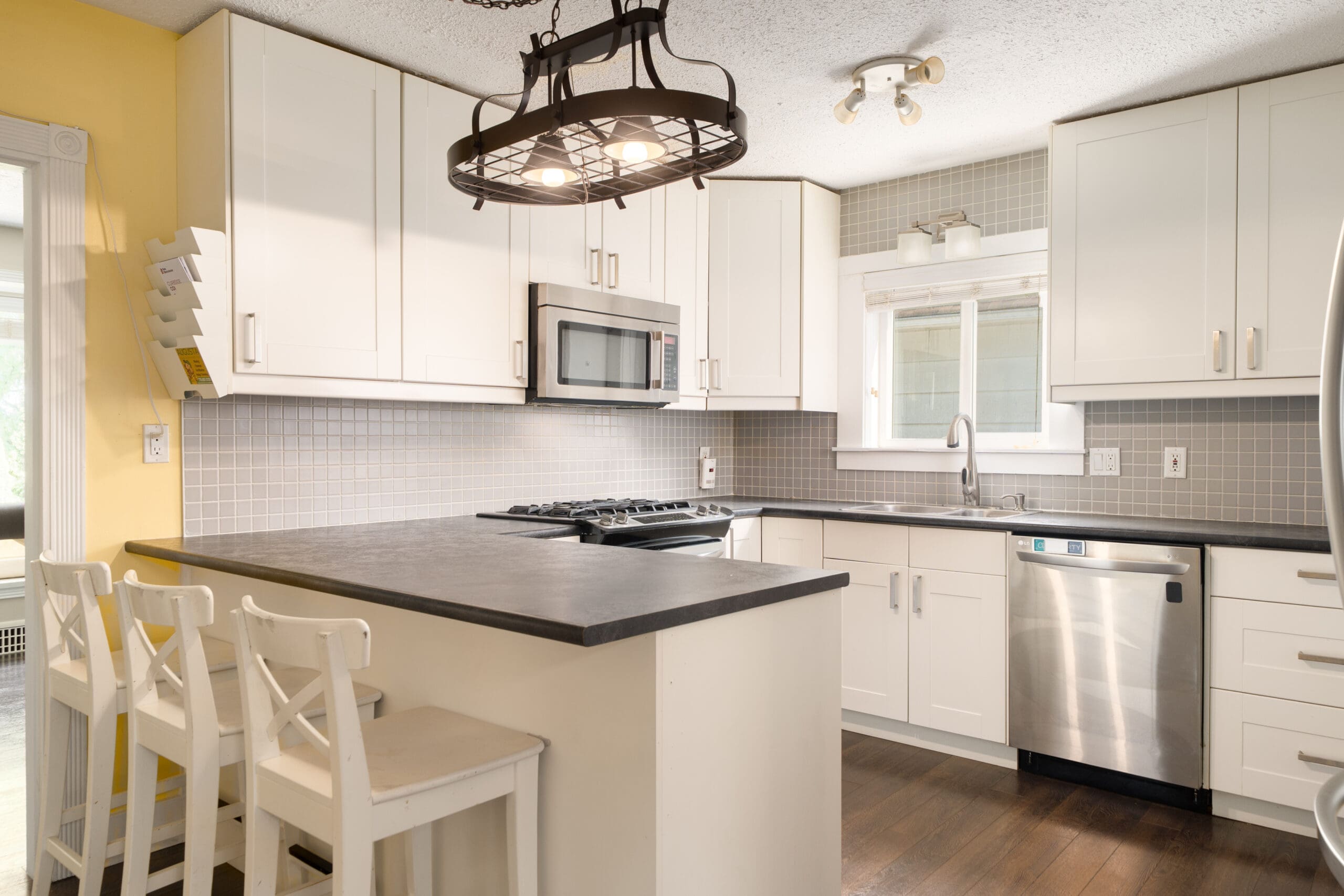 Modern kitchen with white cabinets, stainless steel appliances, gray tile backsplash, black countertop, wooden floor, and three white barstools at a peninsula under a metal light fixture.