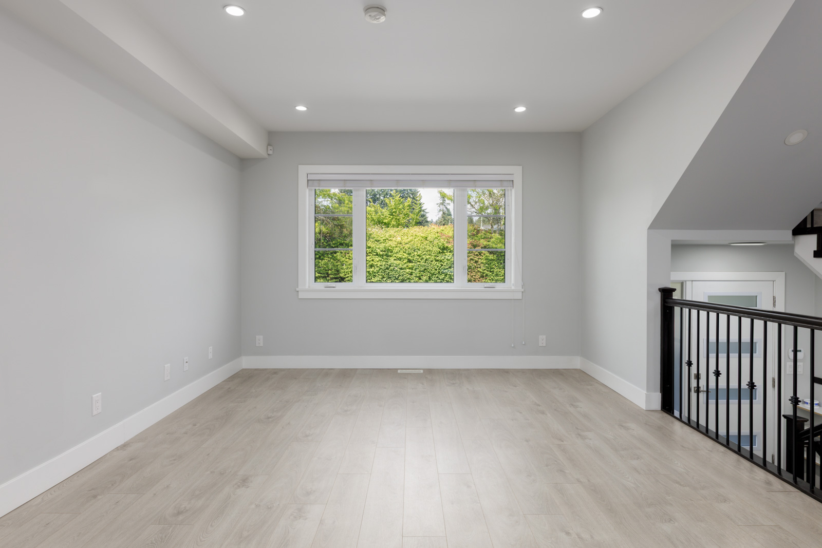 Unfurnished modern townhouse room with light wood floors, white walls, ceiling lights, window view, and black stair railing in Coquitlam.