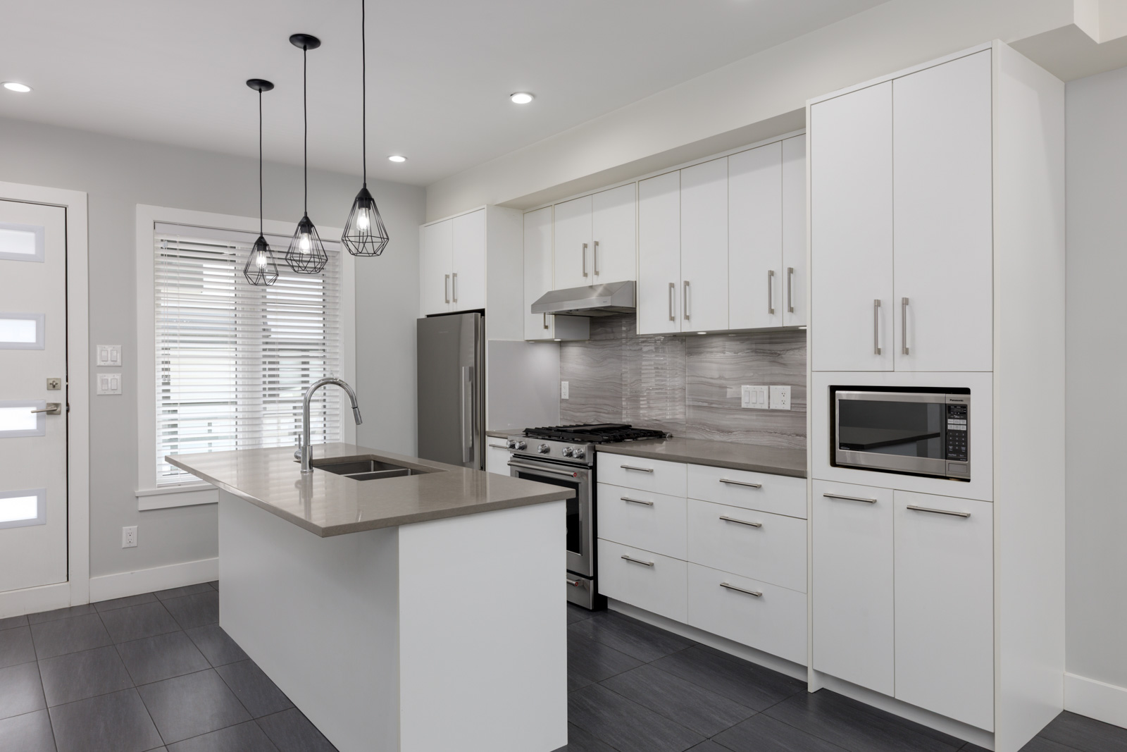 Modern Coquitlam kitchen with white cabinets, stainless appliances, gray backsplash, and island with sink under three pendant lights.