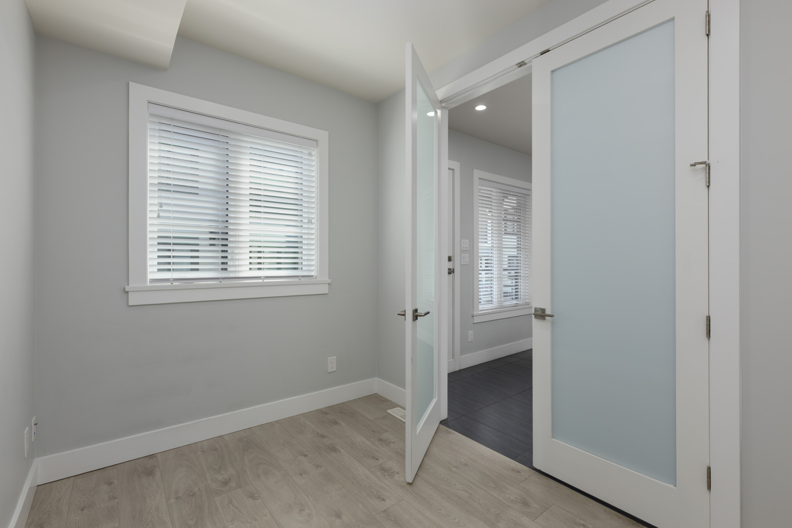Small empty room with light gray walls, white blinds, light wood floor, and an open frosted glass door leading to a dark-tiled hall in Coquitlam.