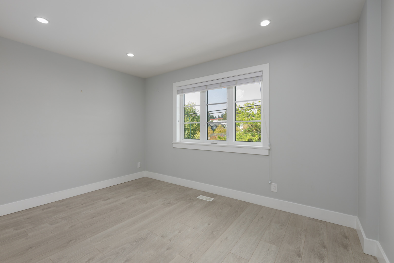 Empty room in a modern Coquitlam townhouse with light gray walls, wood floors, recessed lights, and a large window facing greenery.