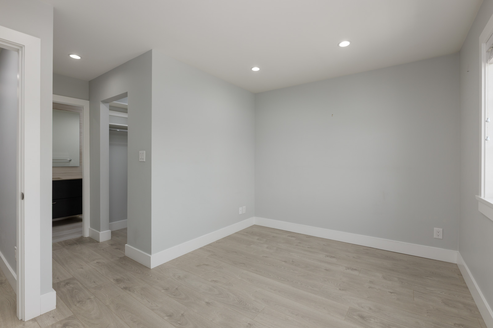 Empty room in a modern Coquitlam townhouse with light gray walls, wood floors, recessed lights, window, closet, and bathroom door.