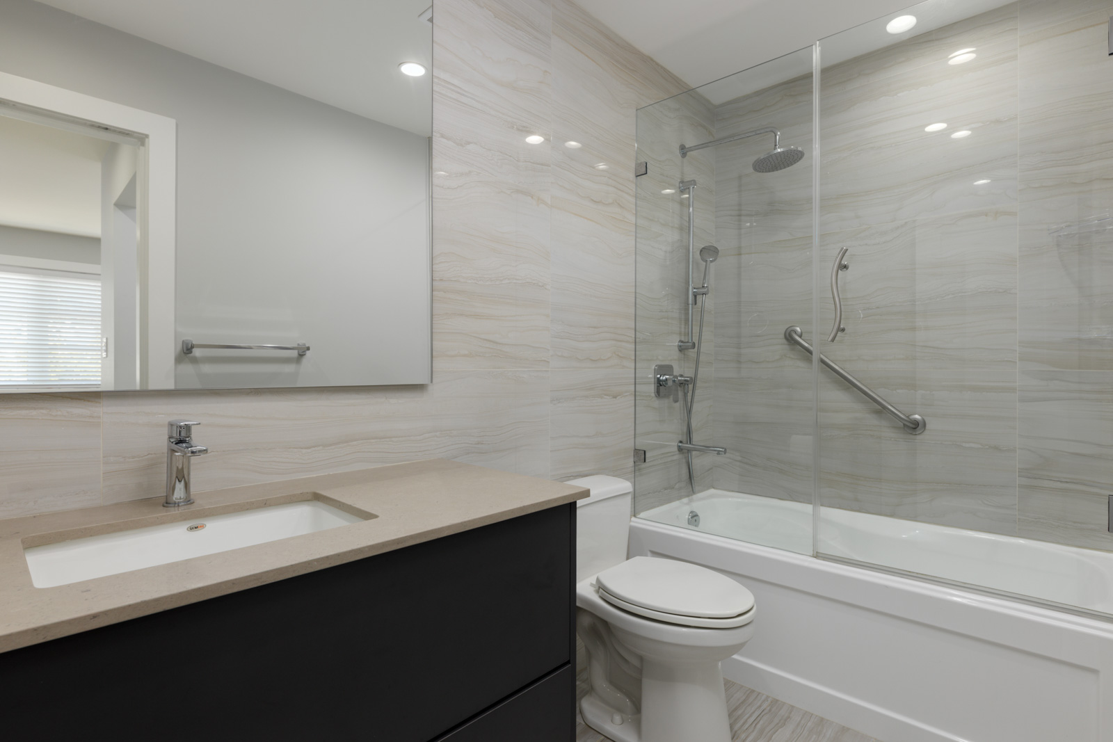 Modern bathroom on Gauthier Ave, Coquitlam with beige countertop, rectangular sink, toilet, glass-enclosed tub, marble walls.