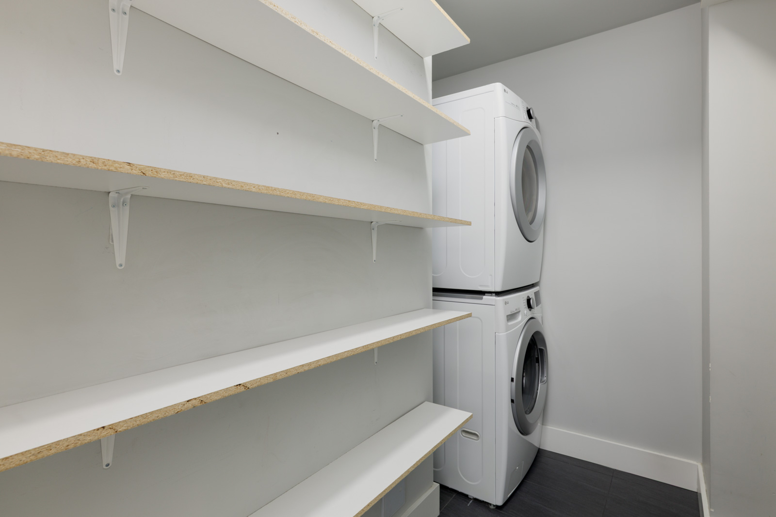 Small Coquitlam laundry room with empty white shelves on the left and a stacked washer-dryer on the right, white walls.