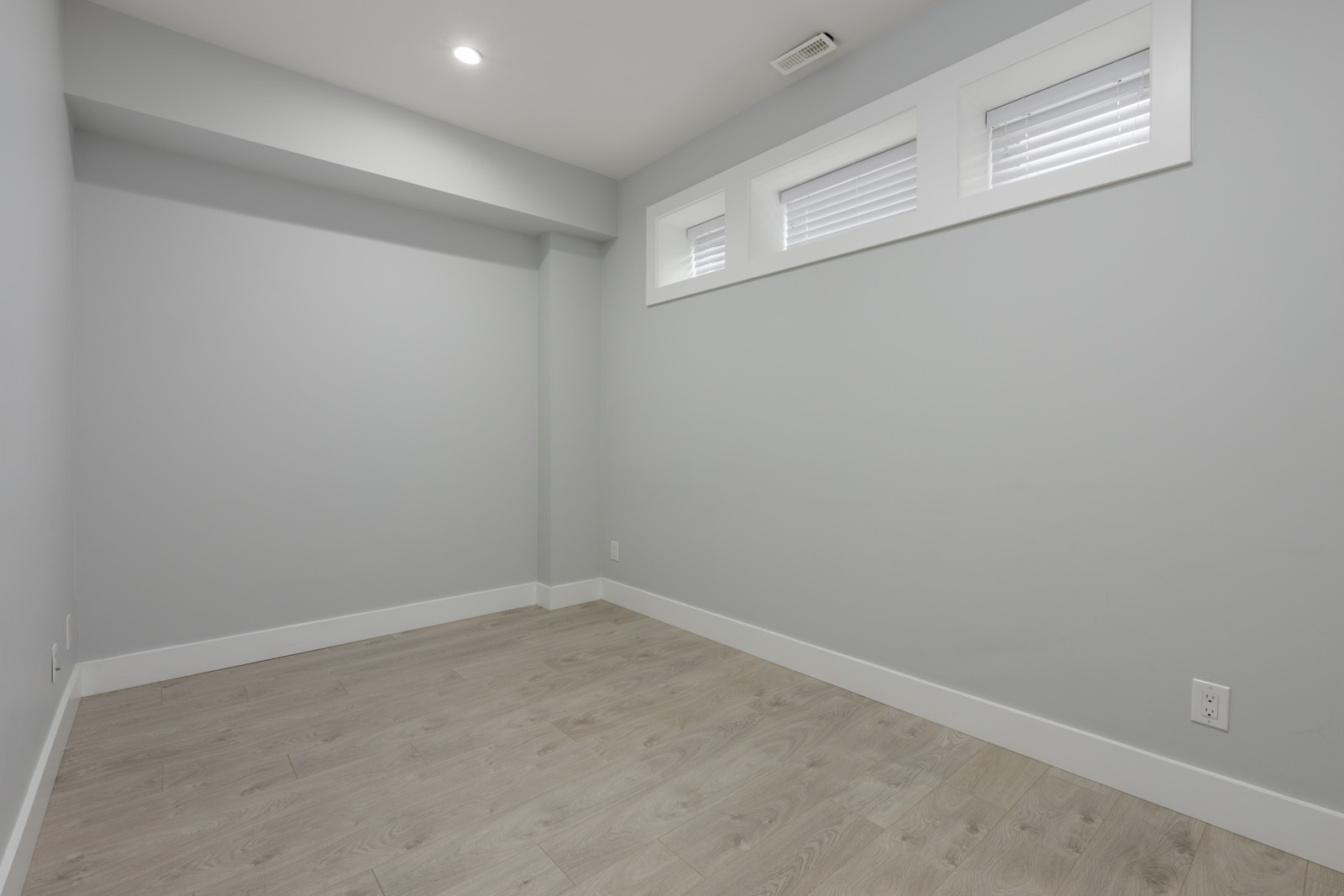 Empty townhouse room with light gray walls, light wood floors, three high rectangular windows, and recessed lights in Coquitlam.
