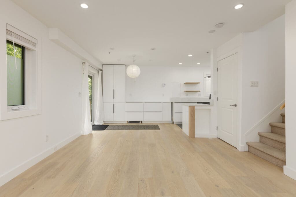 Unfurnished open-plan living area on E 8th Ave, Vancouver with light wood floors, white walls, kitchen, balcony doors, and carpeted stairs.