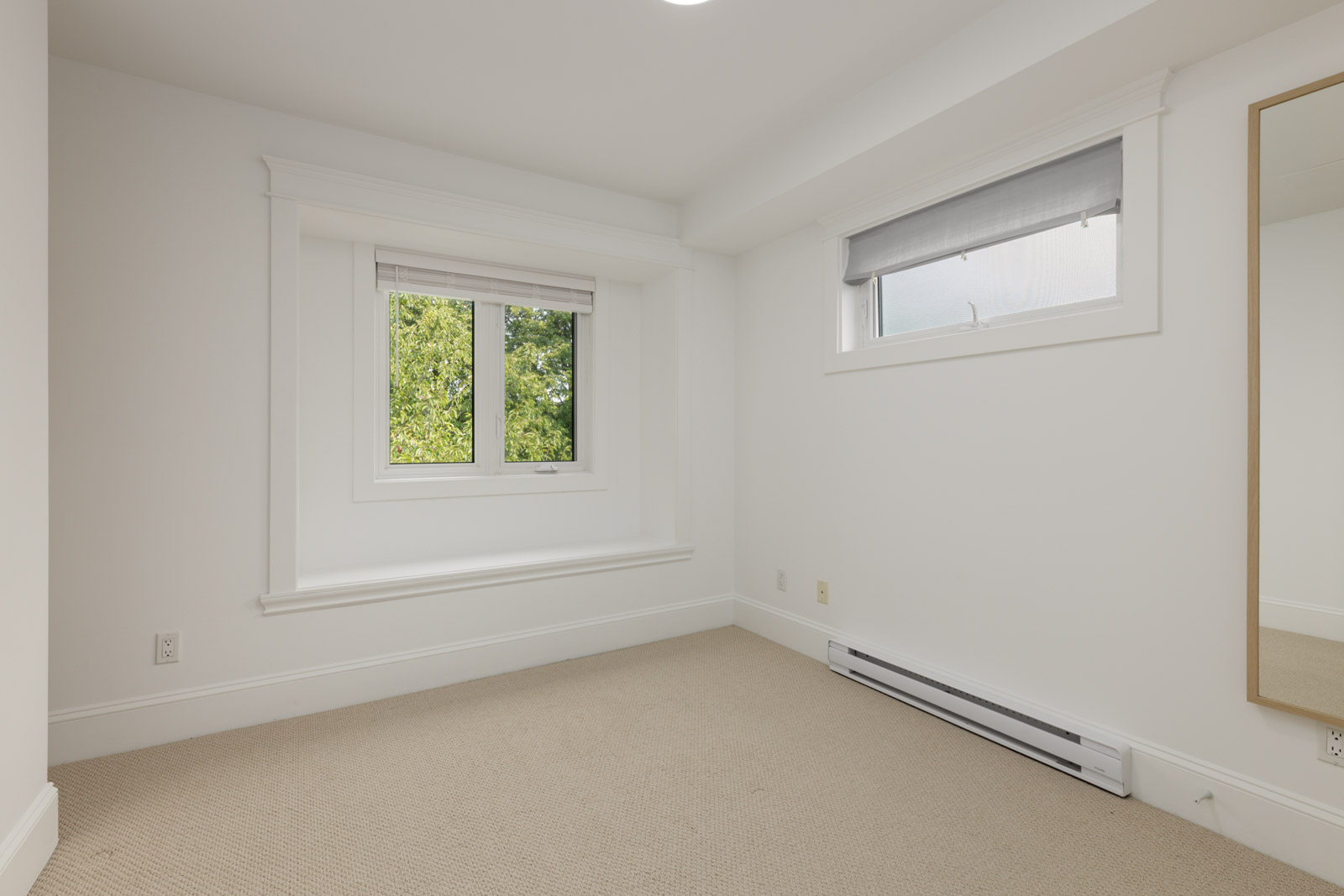 Carpeted, empty room with white walls, two windows with blinds, wall mirror, baseboard heater; green foliage outside. Vancouver.