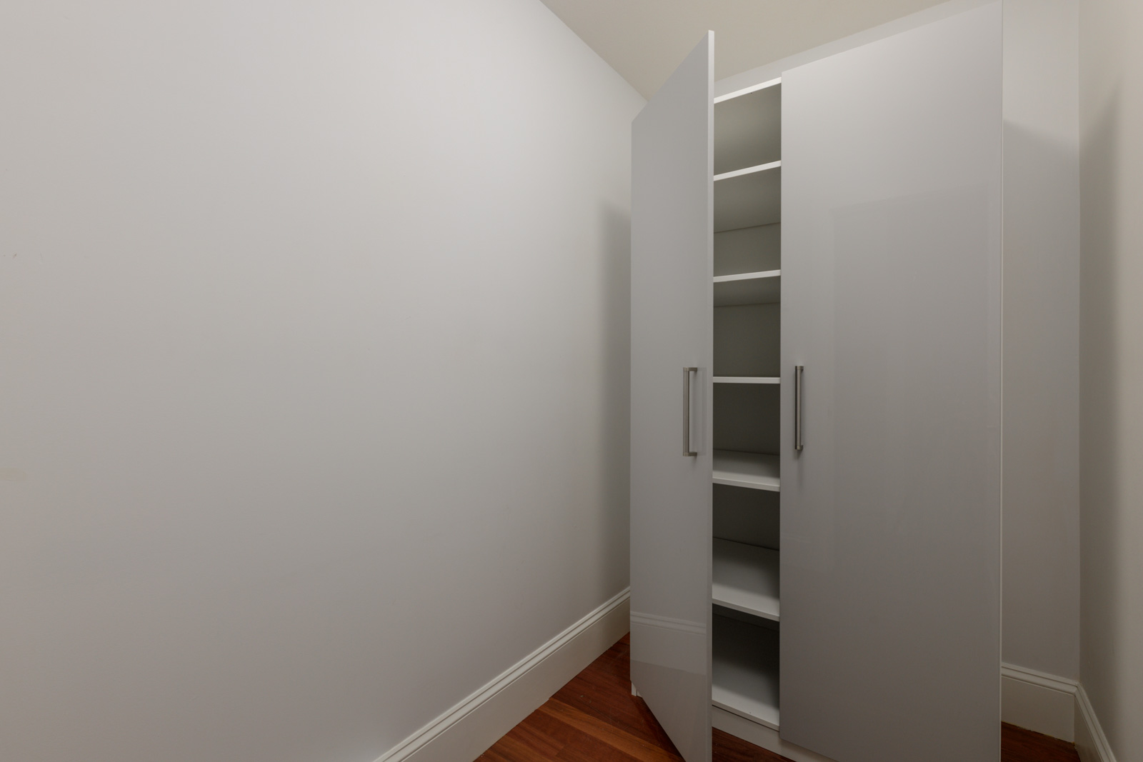A white glossy cabinet with open doors and empty shelves sits by a plain white wall on wood floors, typical of St Catherines St, Vancouver.