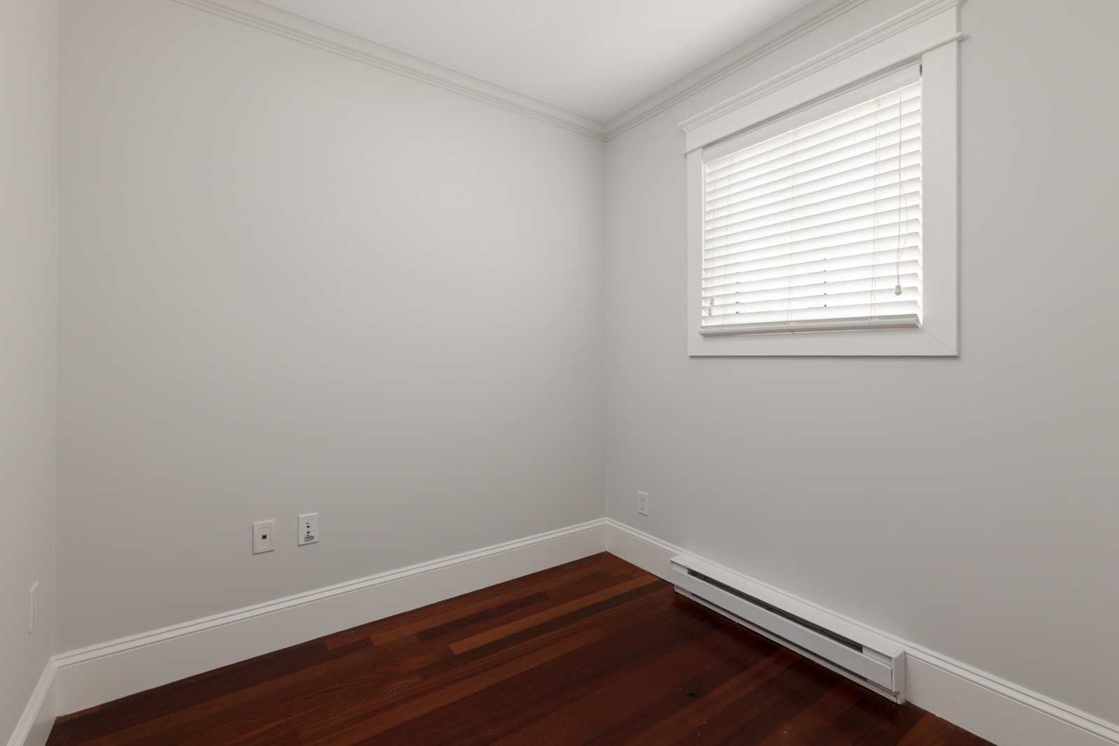 Unfurnished room in Vancouver with white walls, closed-blind window, outlets, baseboard heater, and dark wood floor.