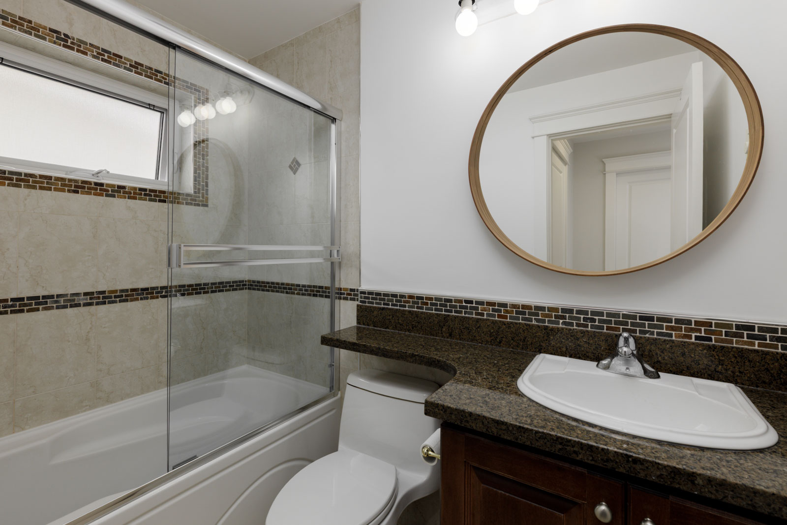 Modern bathroom on E 8th Ave with a bathtub, glass shower doors, toilet, dark granite vanity, sink, and round mirror.