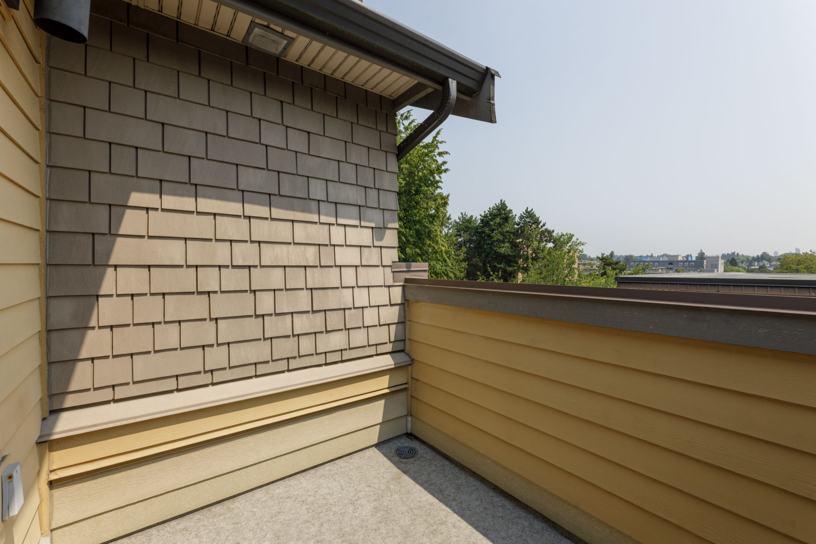Small outdoor balcony with tan siding and shingle wall, concrete floor, overlooking trees and rooftops in Vancouver.