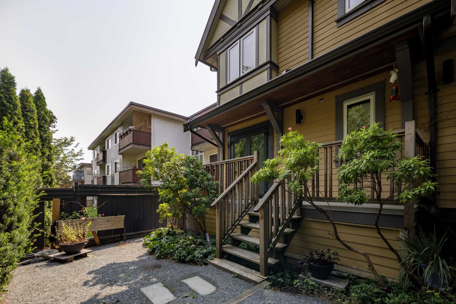 Two-story house with covered porch, wooden steps, and green plants in front on E 8th Ave, Vancouver; gravel path nearby.