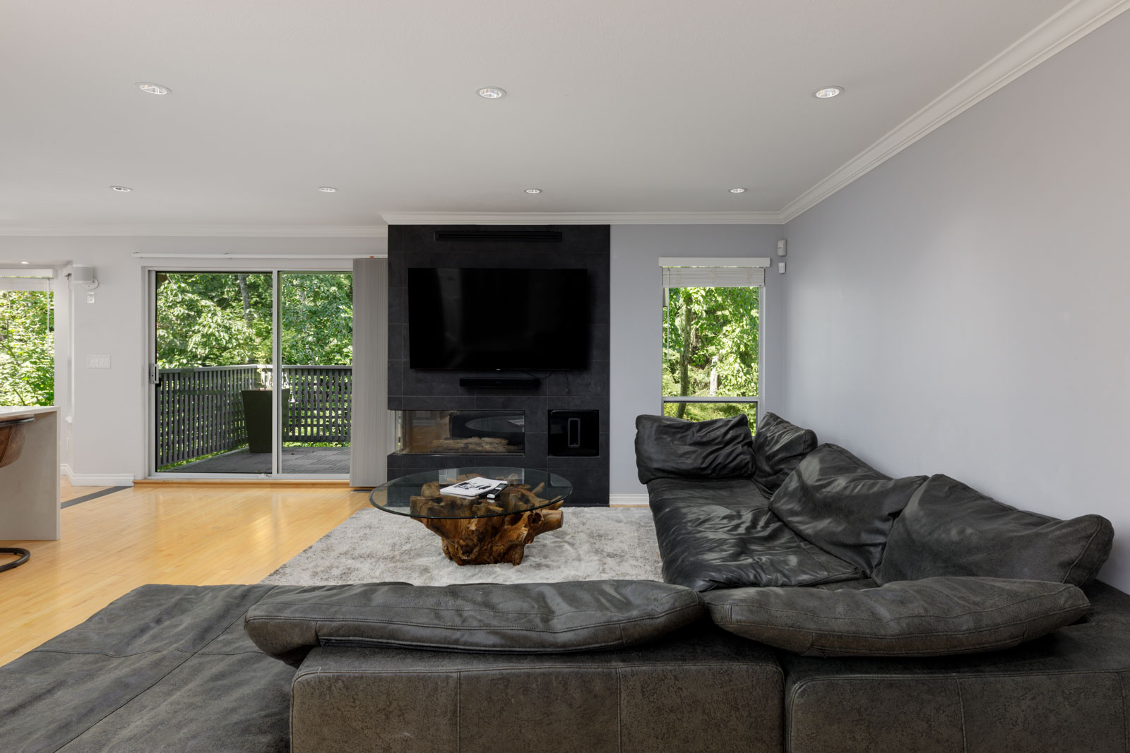 Modern living room in a Vancouver townhouse with black sectional, glass coffee table, wall TV, fireplace, and green view outside.