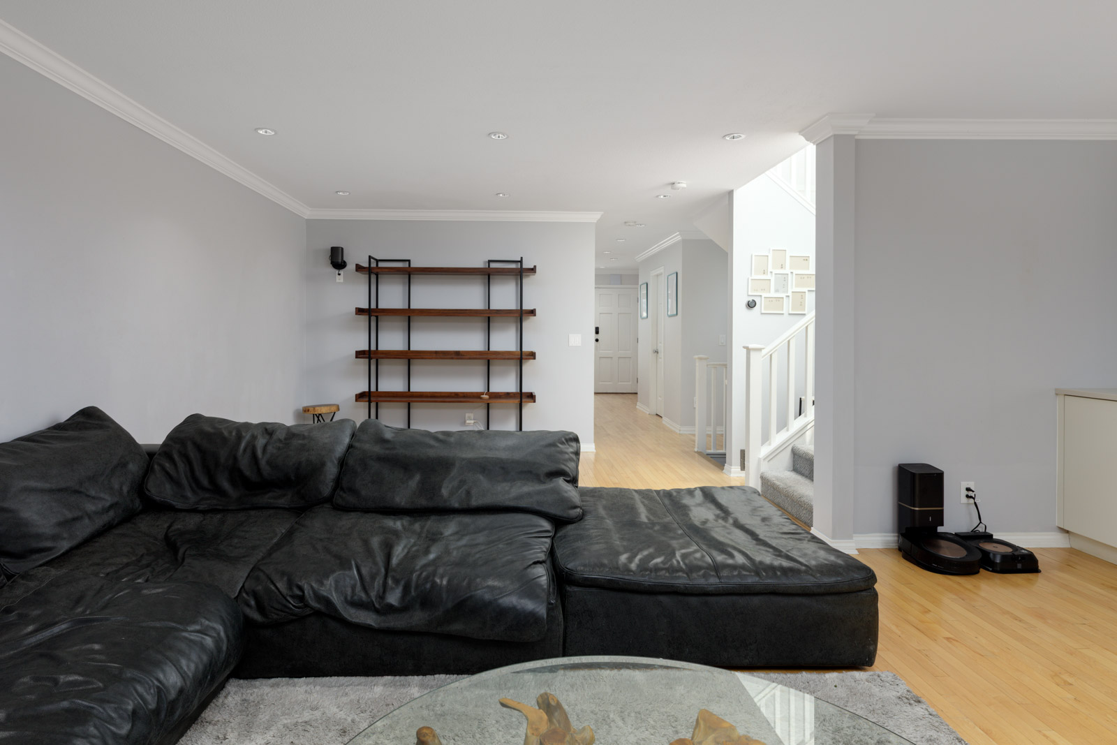 Modern living room in a Vancouver townhouse with a black sectional, glass coffee table, hardwood floor, and robotic vacuum by stairs.