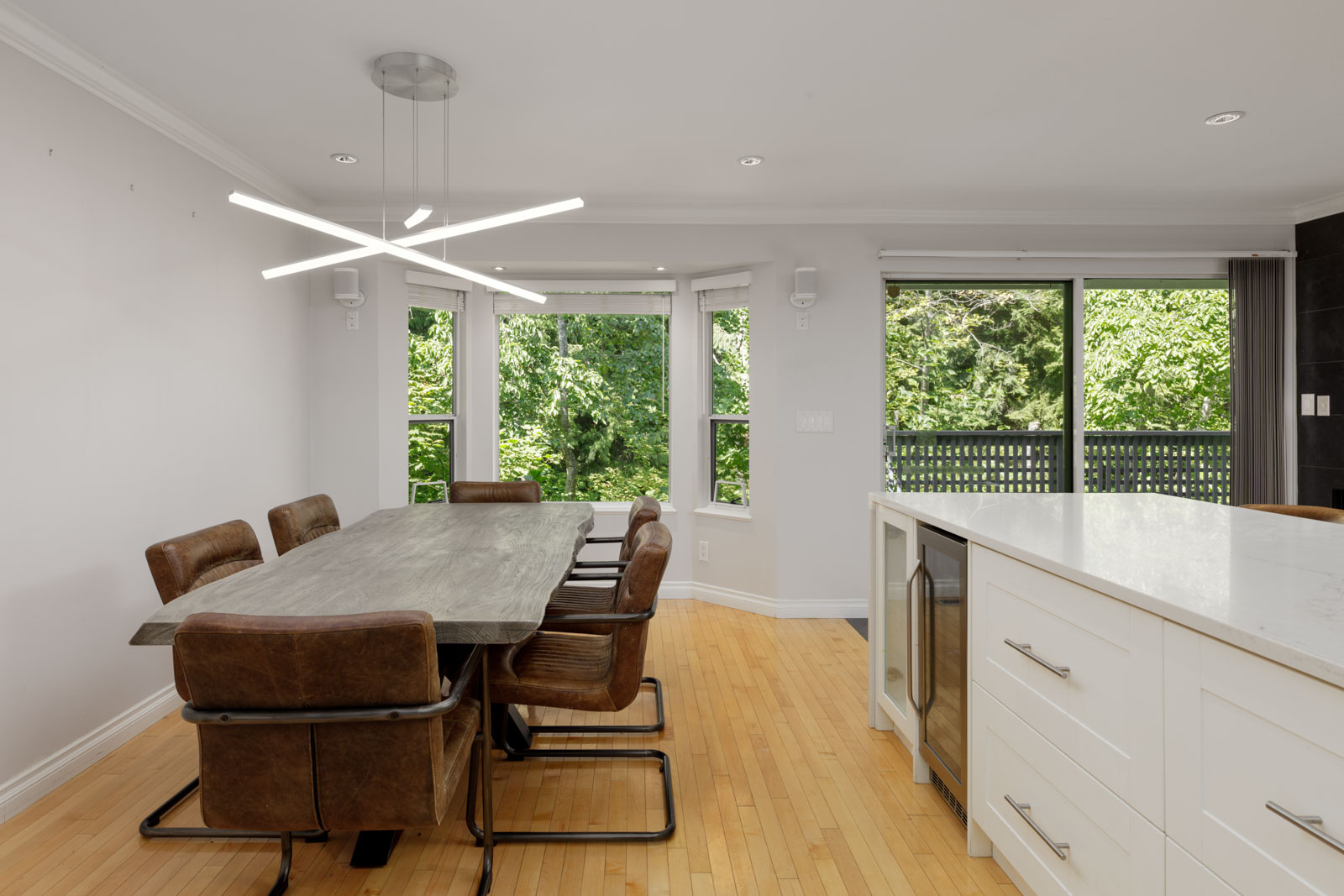 Modern dining area in a Vancouver townhouse with a wooden table, six brown chairs, large windows, and white kitchen island with wine cooler.