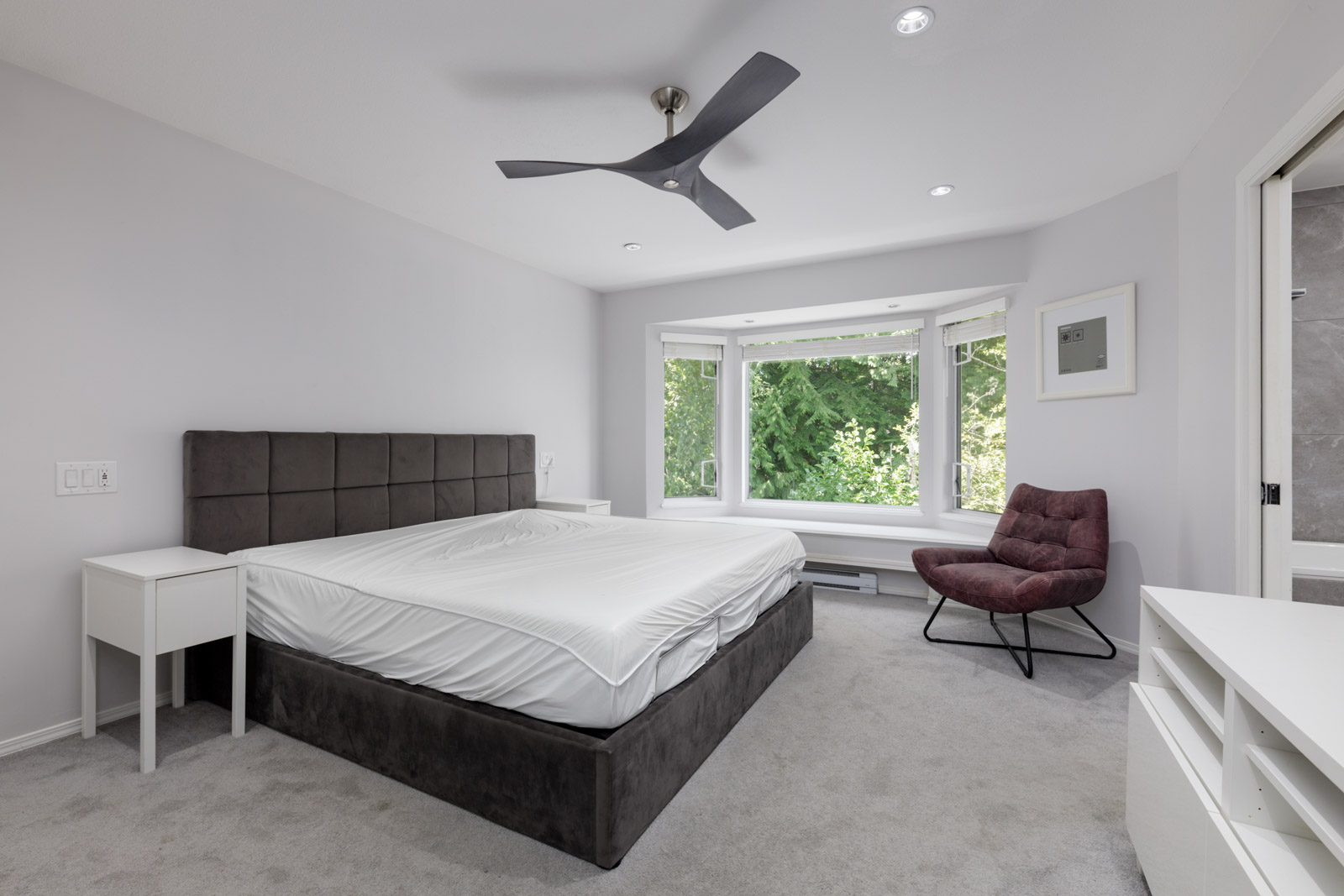 Modern bedroom in a Garibaldi Dr townhouse with a large bed, white nightstands, maroon chair by bay window, and bathroom access.