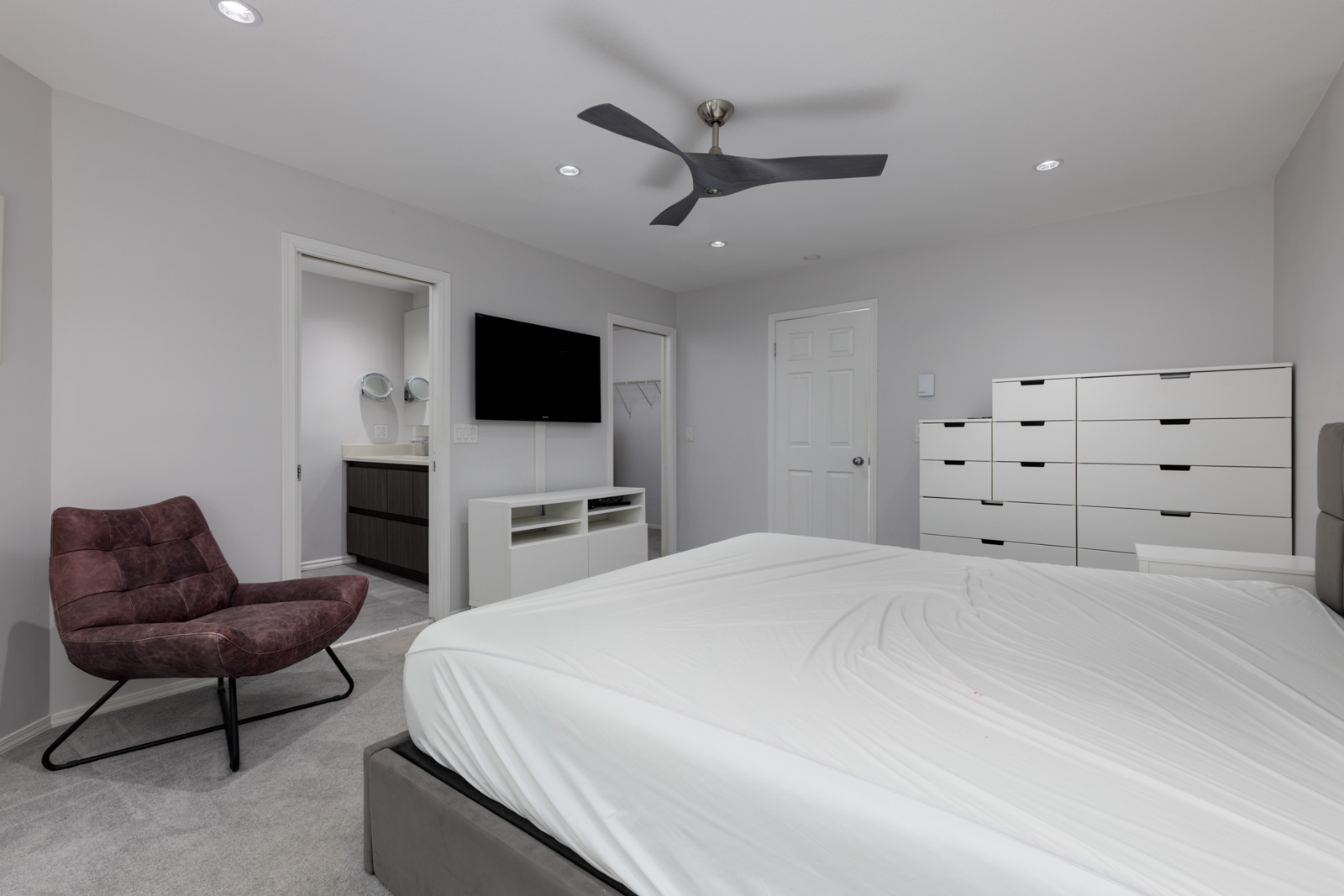 Modern bedroom in a Vancouver townhouse with white dresser, wall TV, purple chair, unmade bed, and bathroom with dual sinks.