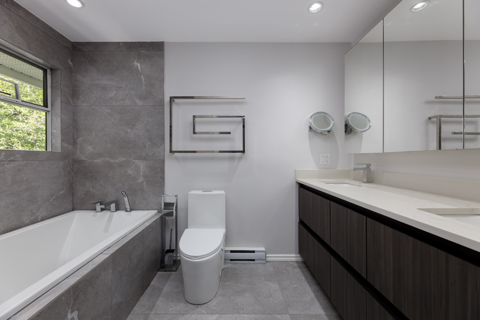 Modern bathroom in a Garibaldi Dr townhouse with gray tile, white tub, wall-mounted toilet, double sink vanity, and window to greenery.