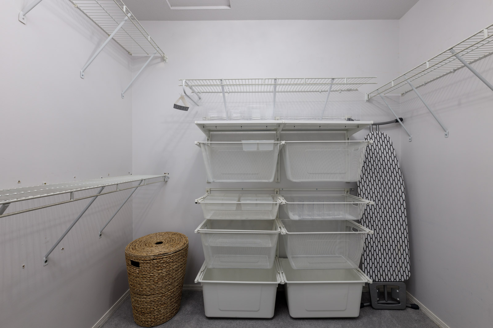 Empty walk-in closet in a Vancouver townhouse with white wire shelves, plastic bins, a wicker basket, and folded ironing board.