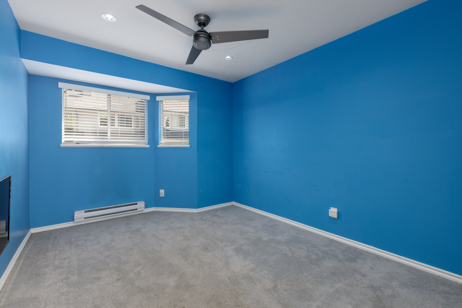 Empty room in a Vancouver townhouse with blue walls, gray carpet, ceiling fan, two windows with blinds, and wall heater.
