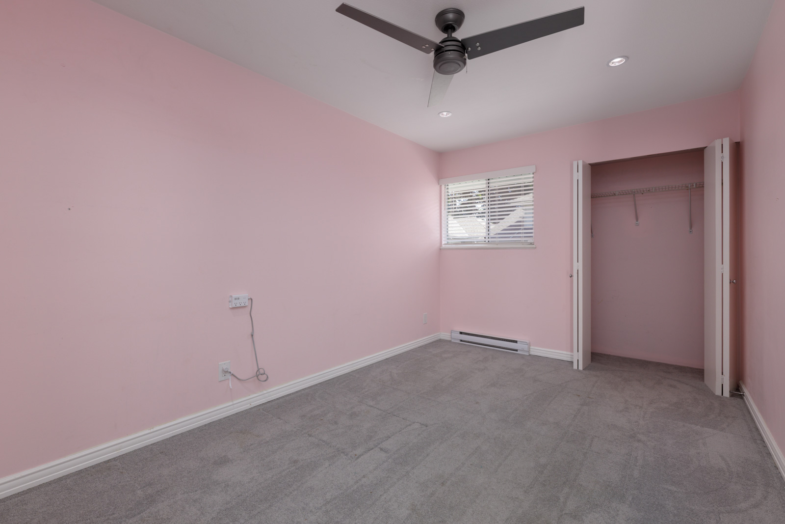 Empty room with pink walls, gray carpet, ceiling fan, small window with blinds, open closet, outlets, and baseboard heater in Vancouver townhouse.