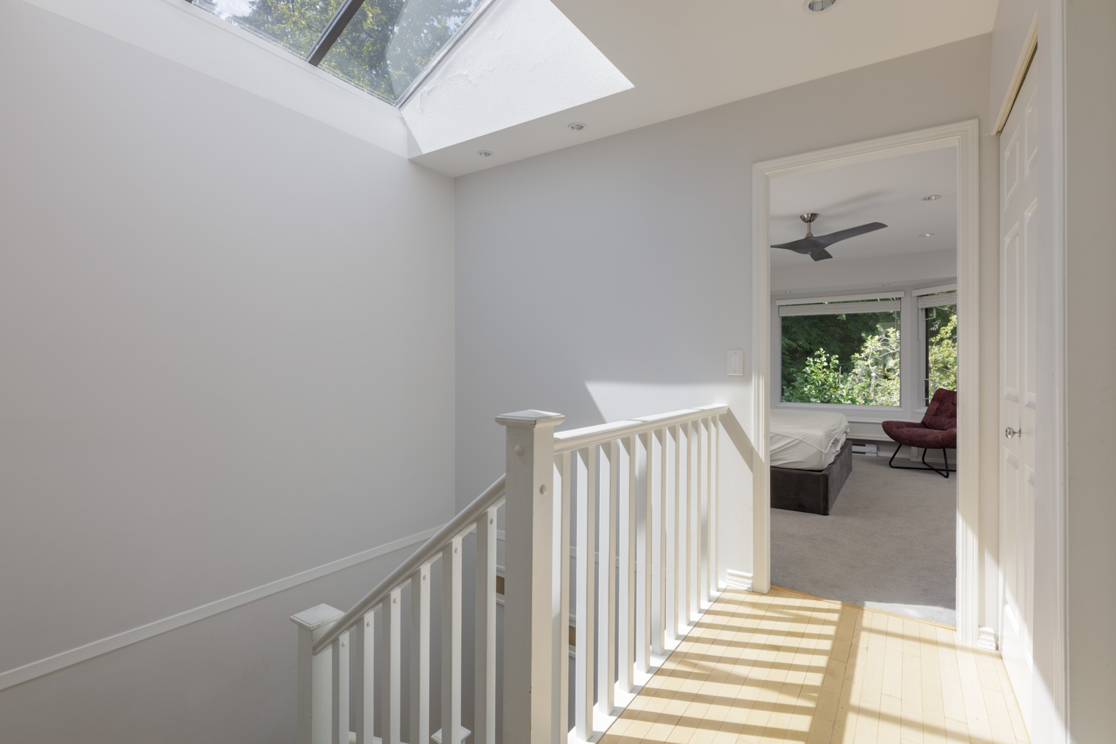 Stairway landing with white railing and skylight in a Vancouver townhouse leads to a bedroom with large windows, fan, and chair.
