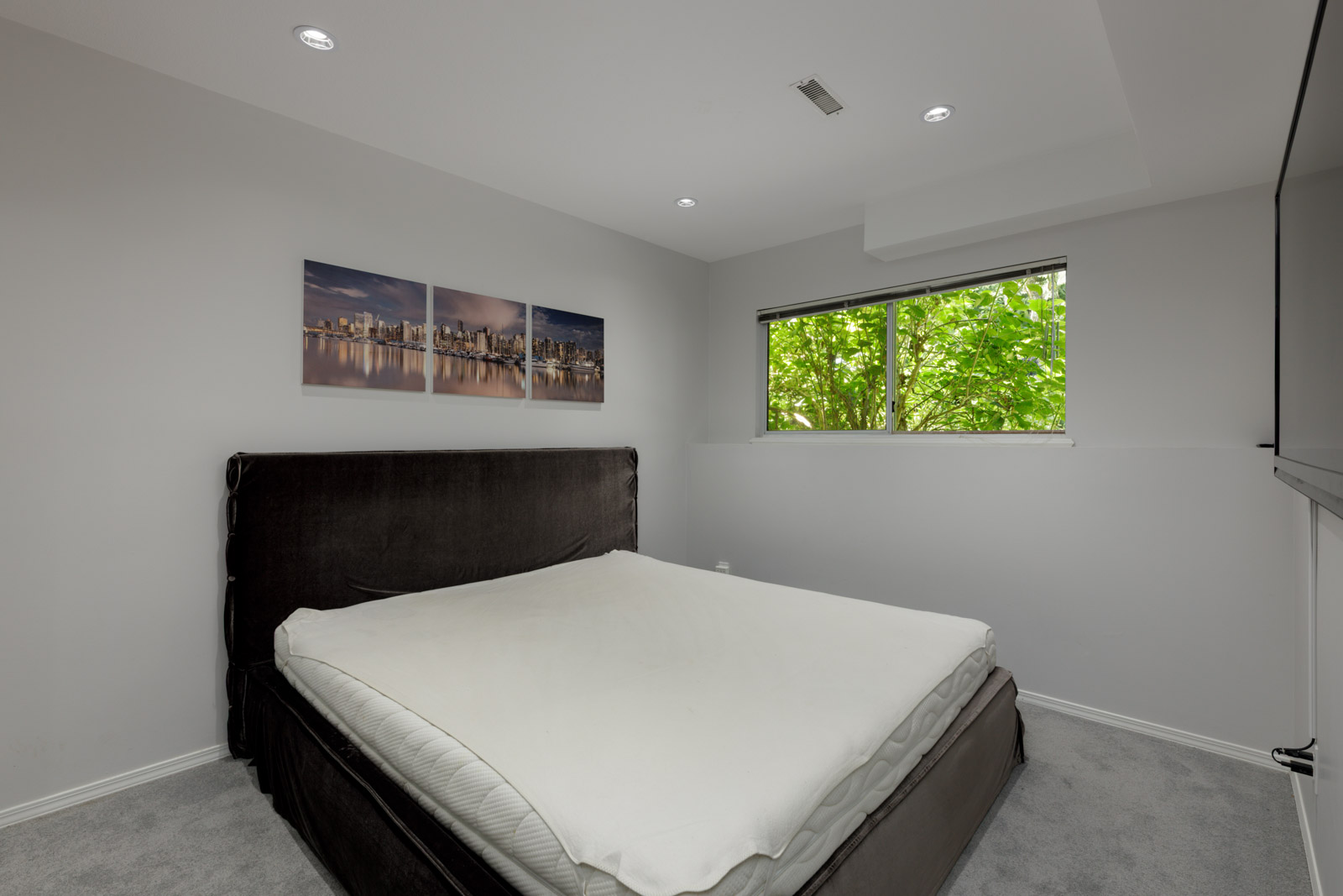 Minimalist Vancouver townhouse bedroom with large dark bed, white bedding, cityscape art, grey carpet, and window facing trees.