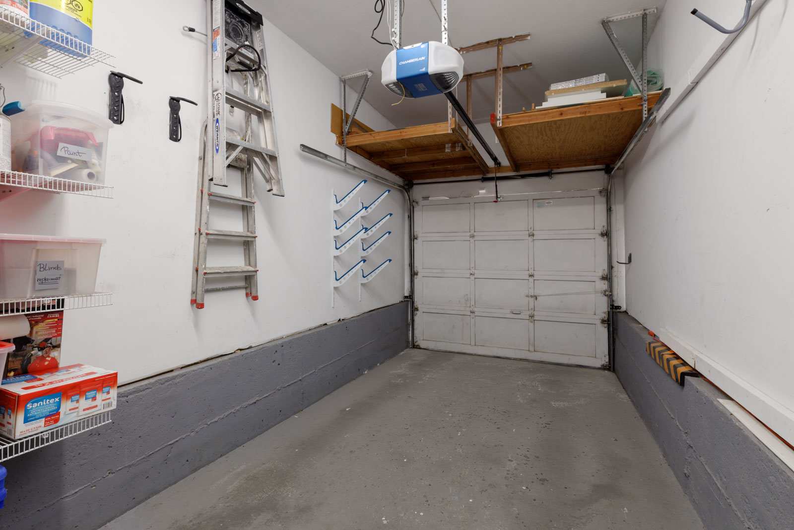 Empty garage in Vancouver townhouse at 3634 Garibaldi Dr with concrete floor, left wall shelves, ladder, hooks, storage, closed door.