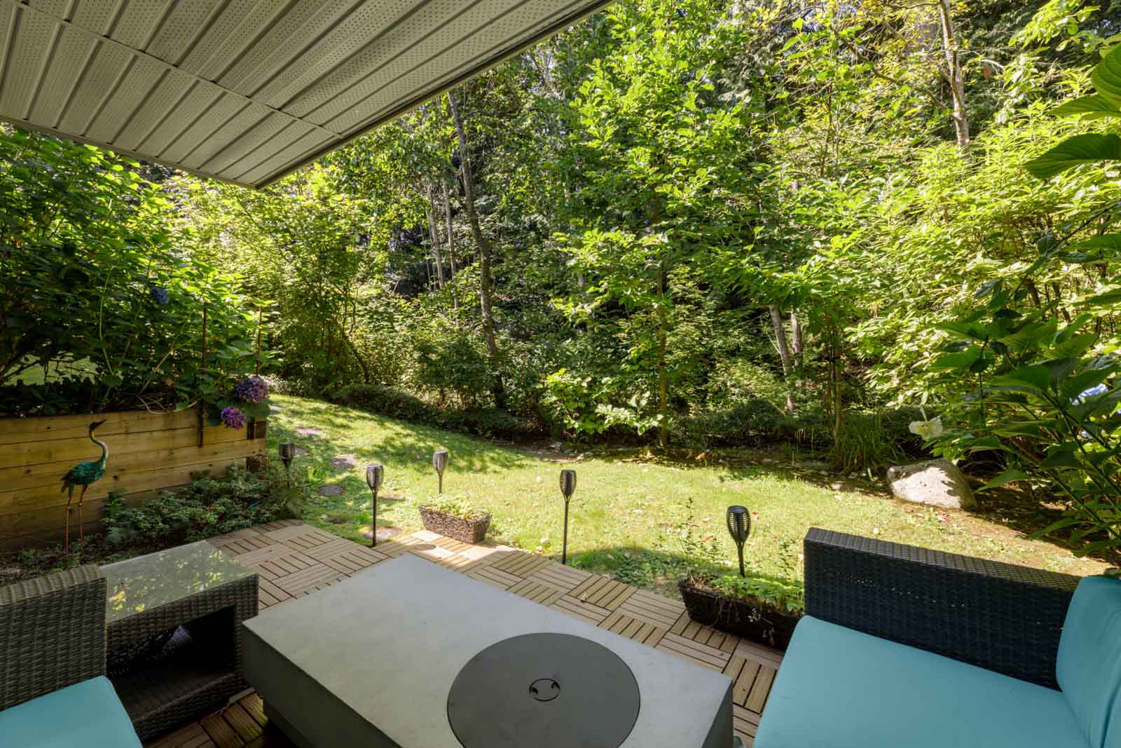 Covered patio with cushioned chairs and a fire pit, overlooking a grassy backyard edged by dense trees in a Vancouver townhouse.