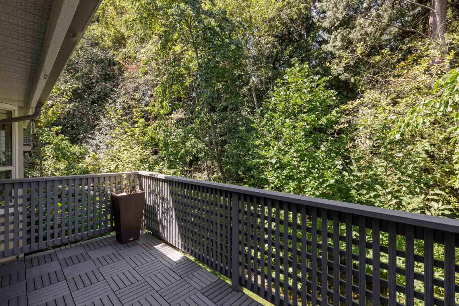 A gray-railed balcony at 3634 Garibaldi Dr, Vancouver, with interlocking tiles and a brown planter overlooks dense green trees.