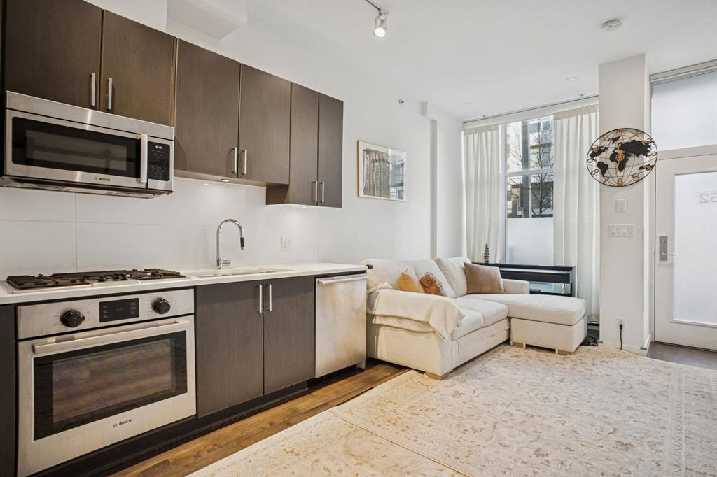 Open kitchen and living space with dark wood cabinets, built-in appliances, white sectional sofa, large windows, and subtle decor.