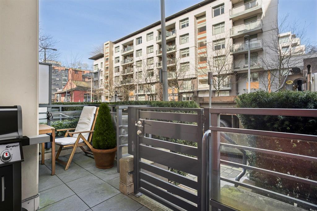 Compact balcony with grill, potted plant, and two chairs, overlooking a street and residential buildings in daylight.