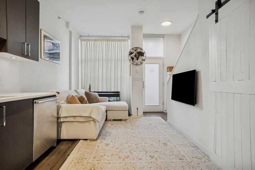 Modern living room with a white sectional, wall-mounted TV, light rug, dark cabinets, and large window with sheer curtains by a door.