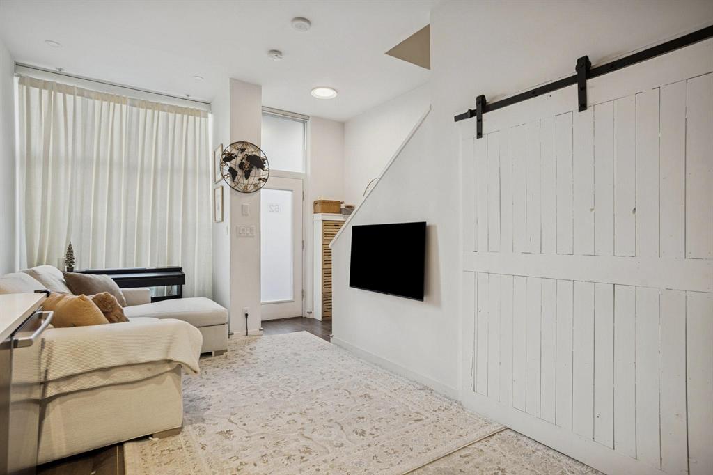 Modern living room featuring a white sectional sofa, wall-mounted TV, large window with sheer curtains, and barn door by the stairs.
