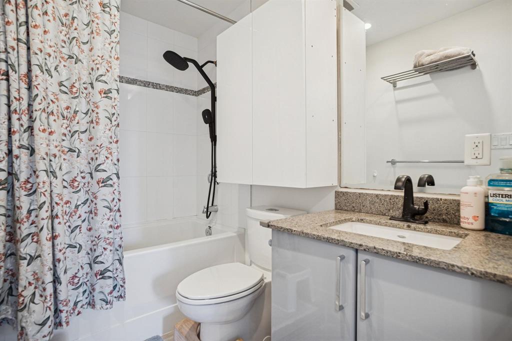Modern bathroom featuring a white tub with floral shower curtain, black showerhead, toilet, granite vanity, mirror, and shelving.