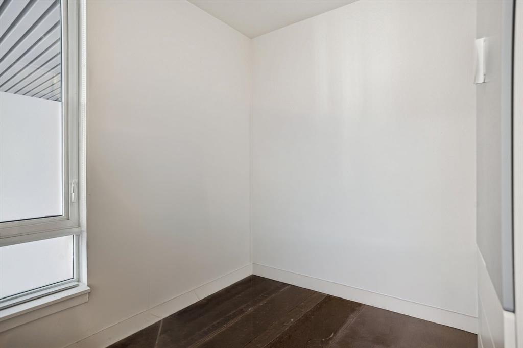 A small, unfurnished room with white walls, a left-side window, and a dark brown floor.