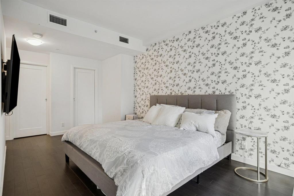 Bedroom featuring a gray upholstered bed, white and gray bedding, floral accent wall, dark wood floor, and round side table.